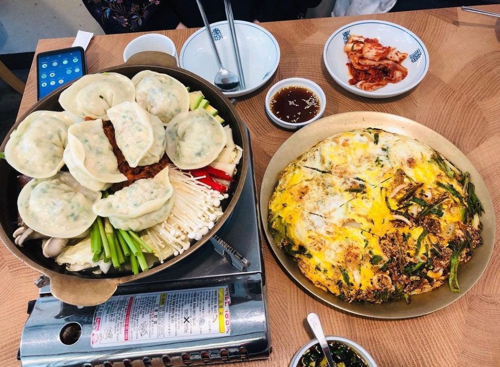A Korean restaurant table with a hot pot filled with handmade dumplings, mushrooms, green onions, and vegetables, served alongside a large golden-brown Korean pancake (jeon), kimchi, and dipping sauces.